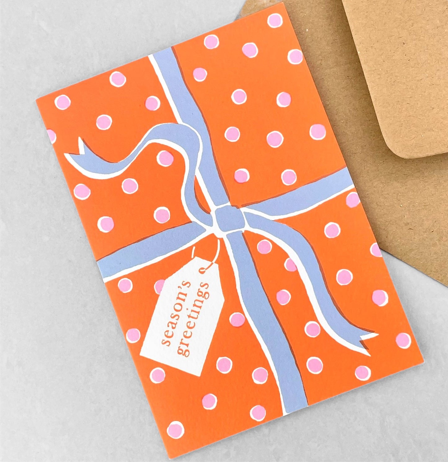 Greetings card with an image of a present wrapped with a bow and a gift tag with "Season's greetings" in orange, pink and blue, by Ariana Martin