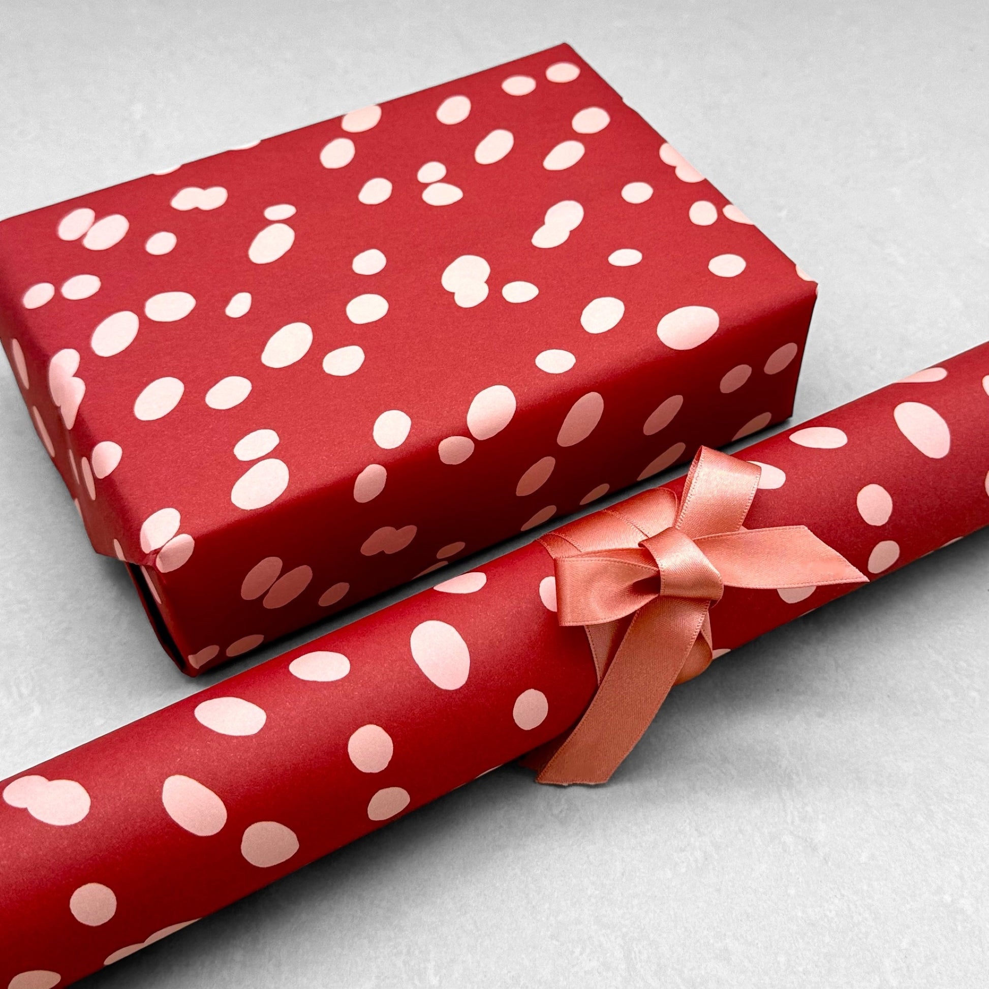 burgundy rouge wrapping paper with random blush pink spots, by Heather Evelyn. Pictured wrapped with a roll of the patterned paper alongside the presente