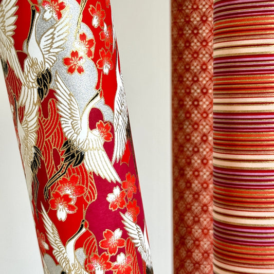 Japanese silkscreen chiyogami paper with a repeat pattern of white cranes in flight on a red and silver background, pictured rolled with other designs