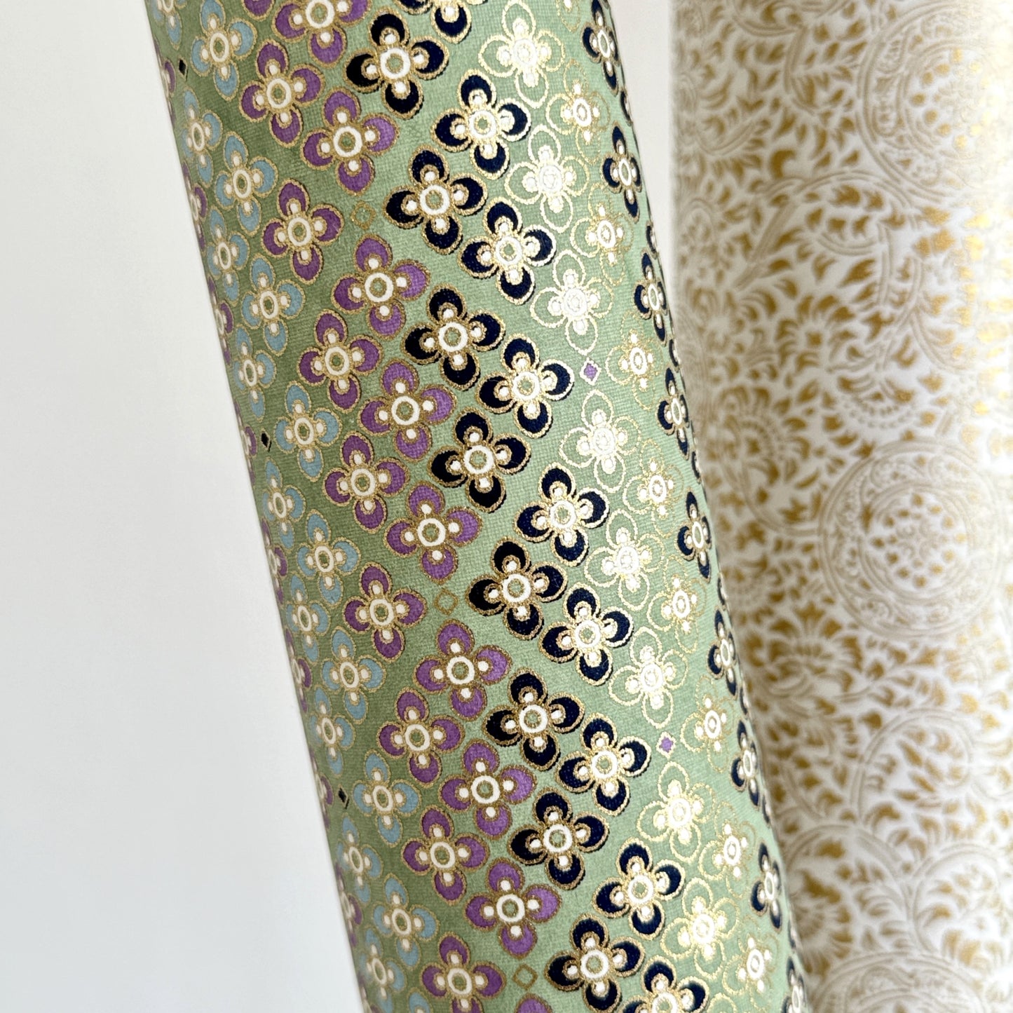 Japanese silkscreen chiyogami paper with a repeat pattern of stripes of a small floral motif in black, lilac and blue on a moss green backdrop, close up rolled
