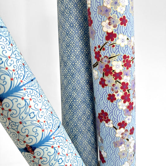Japanese silkscreen chiyogami paper with a pattern of deep red, lilac and white plum blossom on a blue backdrop, rolled and alongside other designs