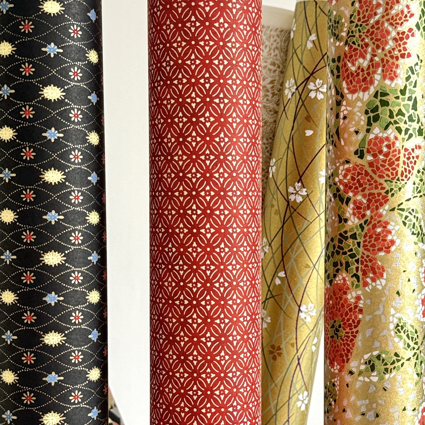 Japanese silkscreen chiyogami paper with a pattern of small repeat motifs in red, blue, lemon and gold on a black background, pictured rolled alongside coordinating designs