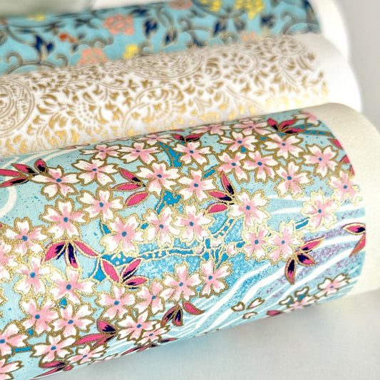 Japanese silkscreen chiyogami paper with an abundance of pale pink cherry blossom with a pale blue backdrop of meandering water, finished with metallic gold ink. Close-up