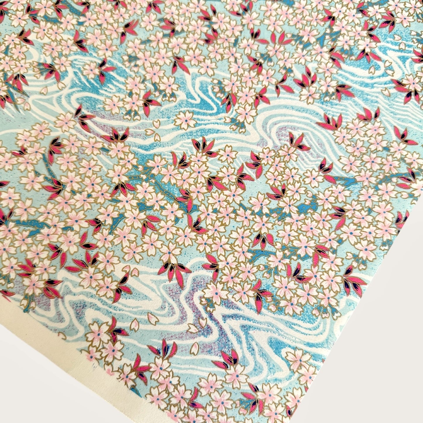 Japanese silkscreen chiyogami paper with an abundance of pale pink cherry blossom with a pale blue backdrop of meandering water, finished with metallic gold ink.