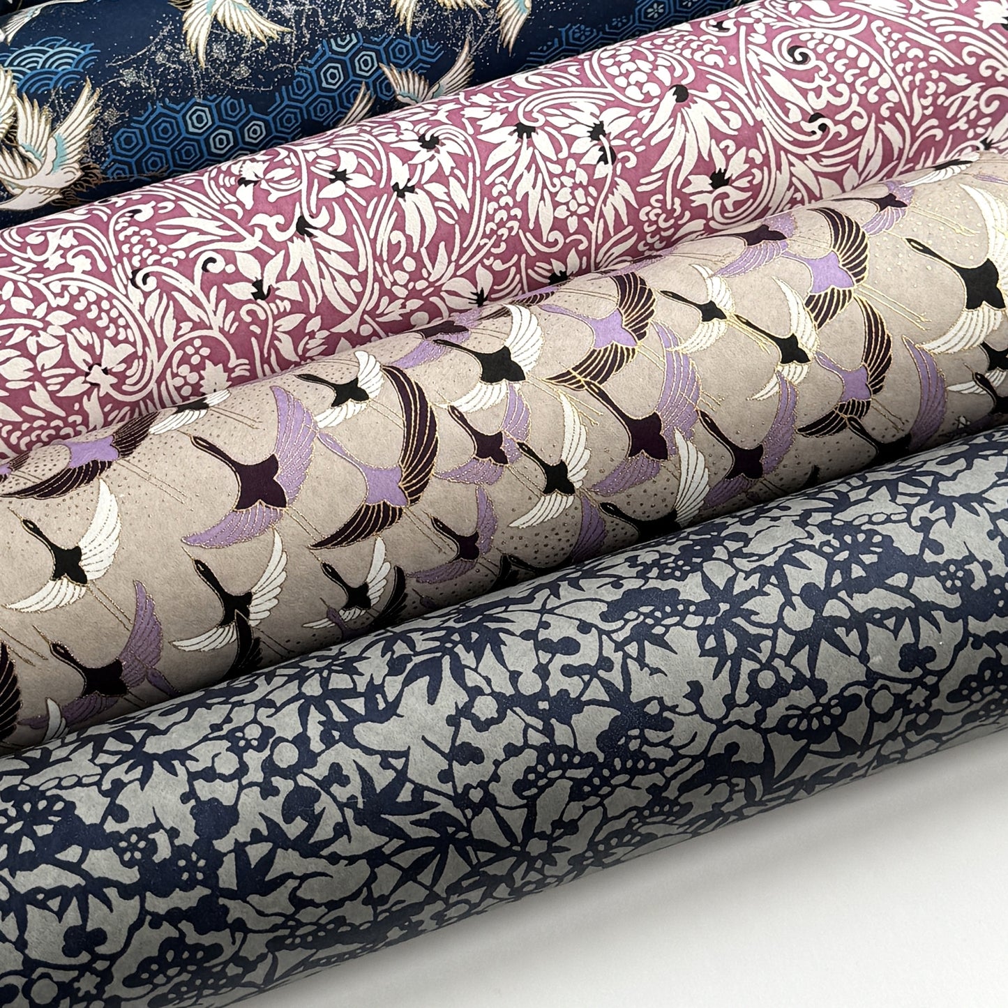 a sheet of Japanese chiyogami yuzen paper with a pattern of cranes in flight in lilac, black, taupe and gold. Pictured with other silkscreen paper designs