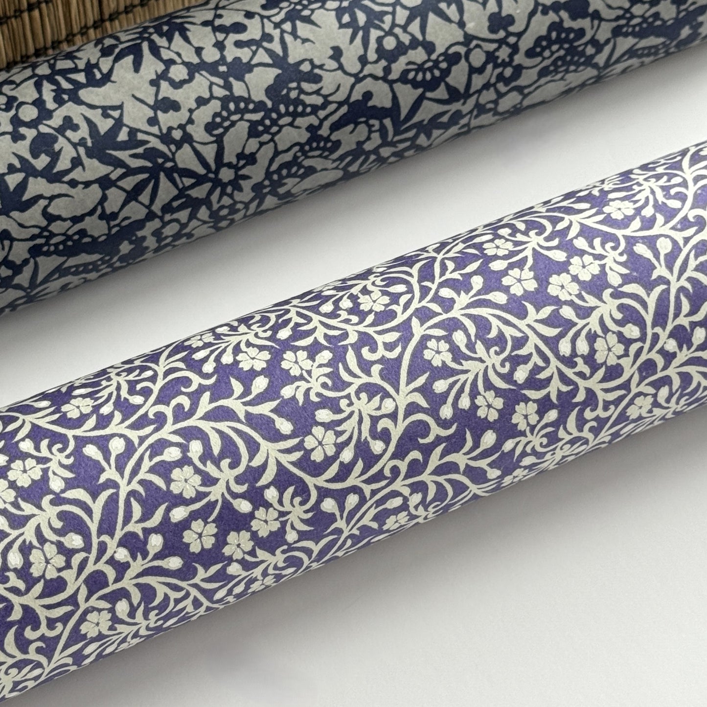 a sheet of Japanese silkscreen printed chiyogami paper with a small floral repeat pattern in grey and white on a periwinkle blue background. Pictured rolled