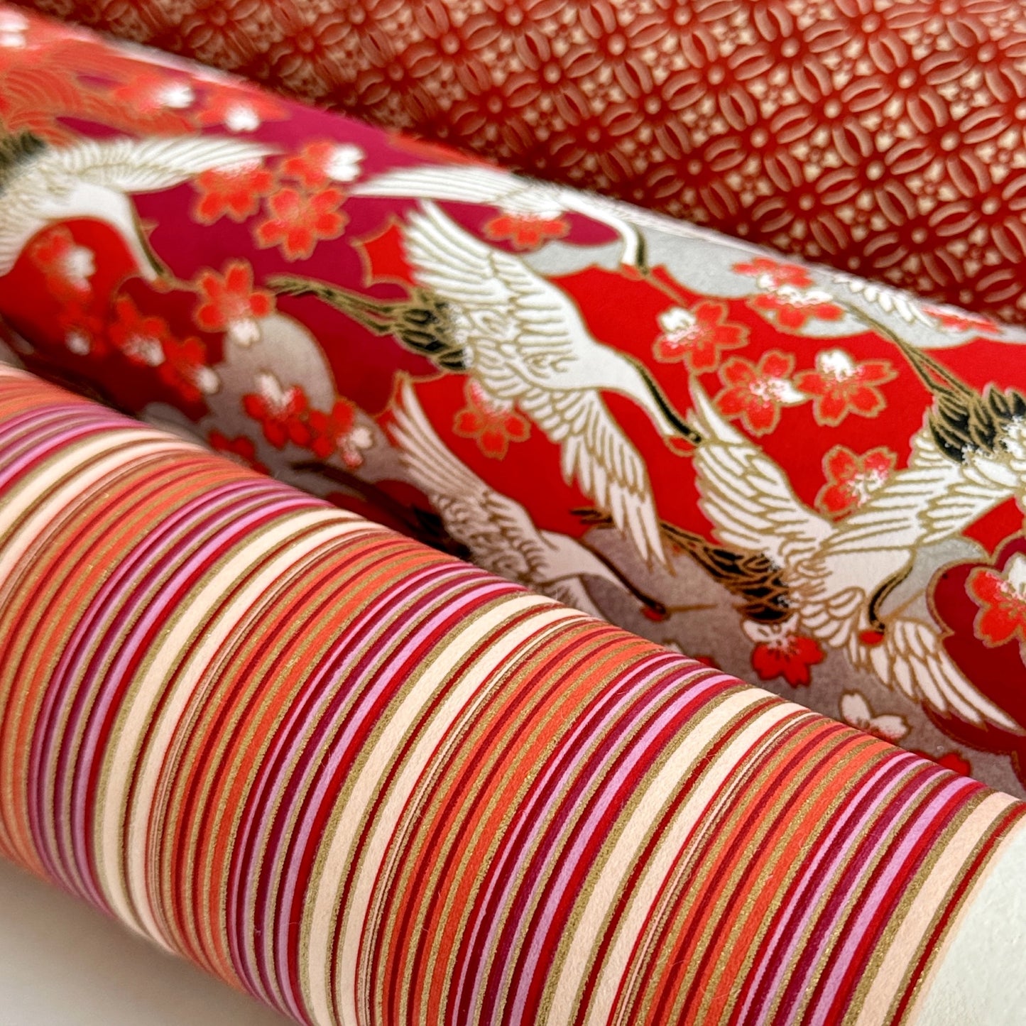 Japanese silkscreen chiyogami paper with a classic narrow stripe pattern in berry tones of red, pink, purple and gold. Pictured rolled alongside other designs