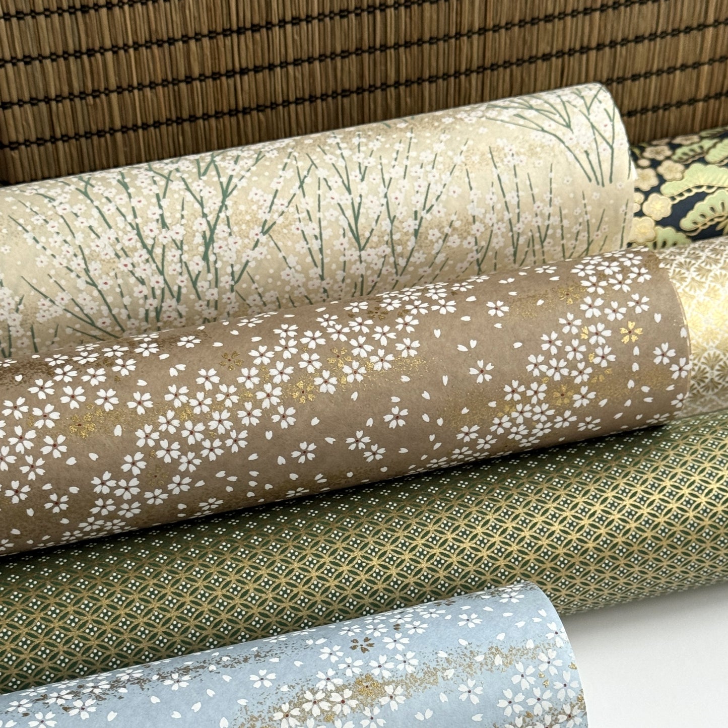 a sheet of Japanese silkscreen printed chiyogami paper with a delicate design of tufts of meadow grasses in flower in white, green on a cream background. Pictured rolled with other designs