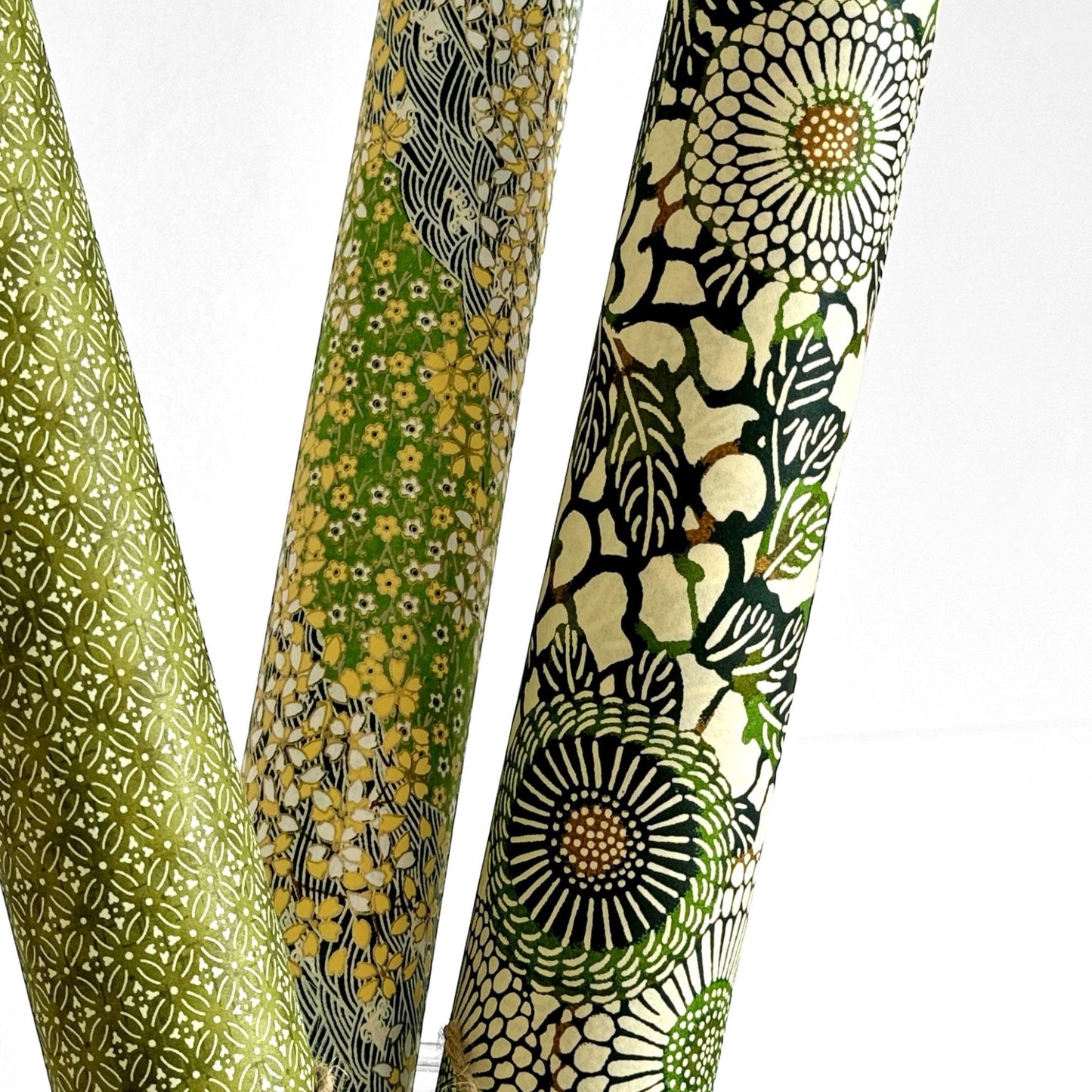 Japanese silkscreen chiyogami paper with a repeat pattern of stylised chrysanthemum flowers in tones of deep green on a buttermilk cream backdrop. Pictured rolled with other designs