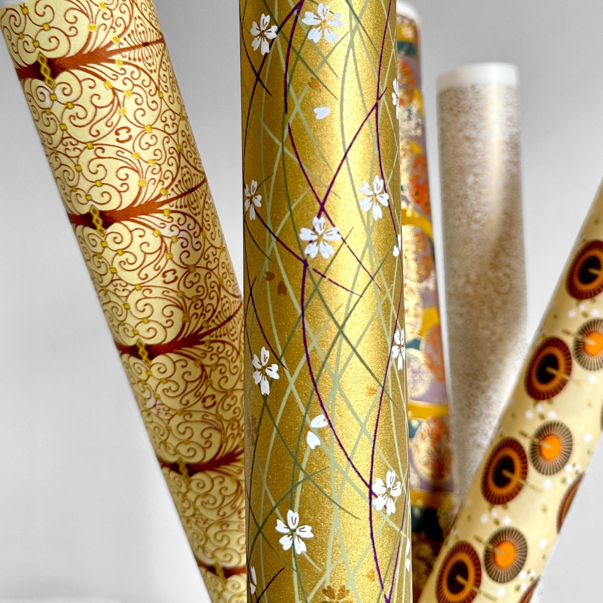 Japanese silkscreen chiyogami paper with a pattern of purple arches and white cherry blossom on antique gold backdrop. Rolled alongside other paper designs