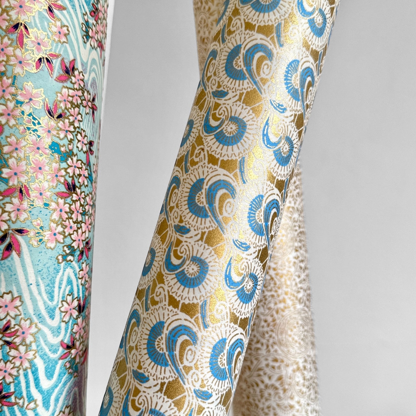 Japanese silkscreen chiyogami paper with a repeat floral pattern in gold an cerulean blue, pictured rolled with other designs