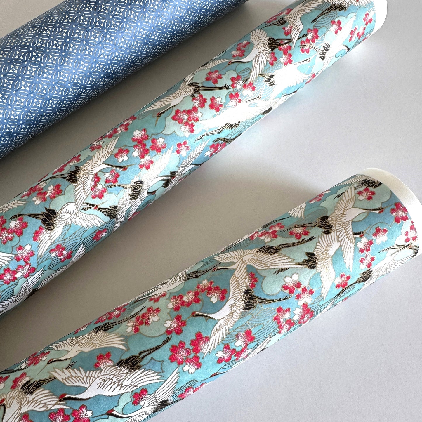 Japanese chiyogami paper with a pattern of white cranes, pink cherry blossom on a blue background. Rolled