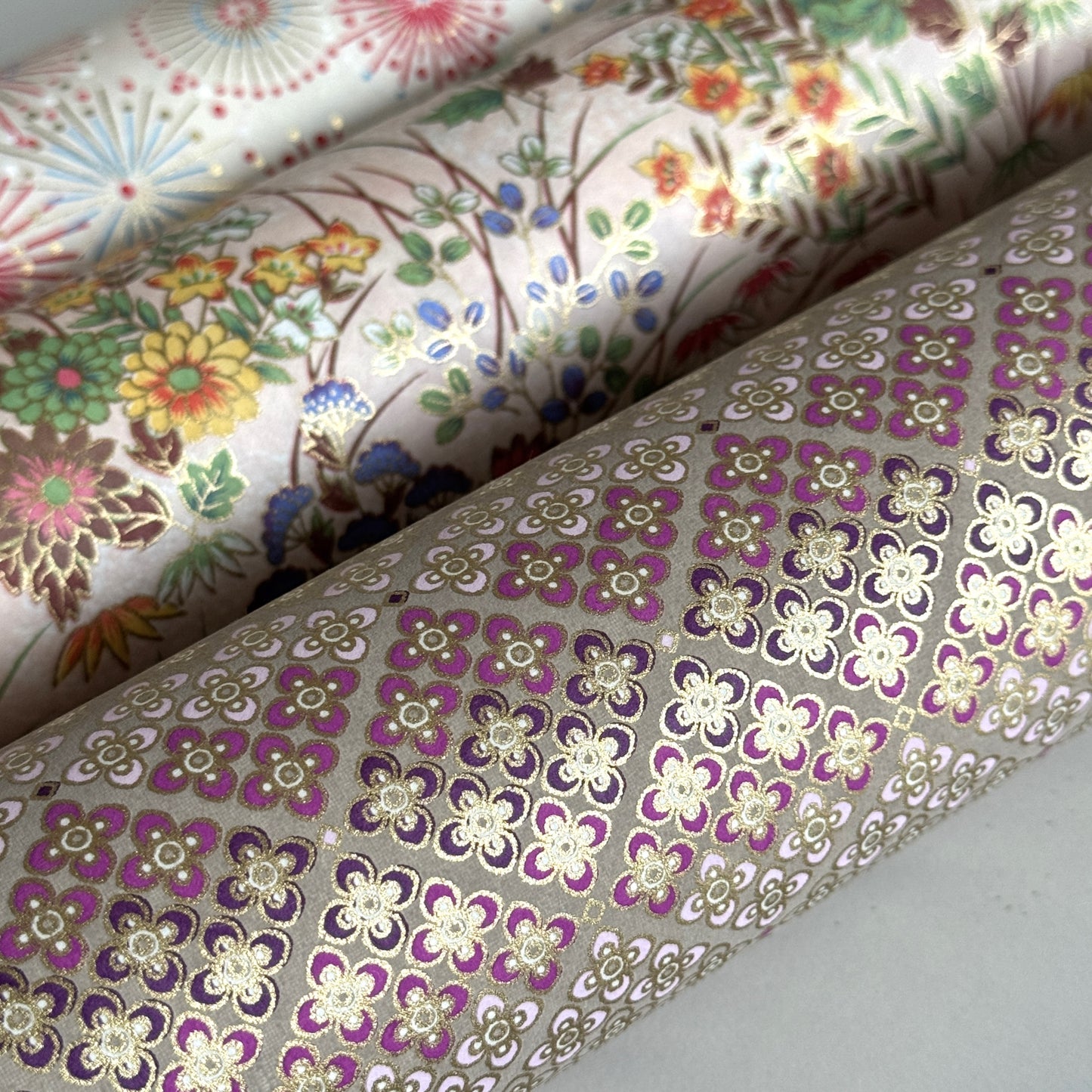 Japanese chiyogami silkscreen printed paper with rows of small flower motifs in purple, lilac and white on a taupe background with gold accents. Rolled alongside other papers