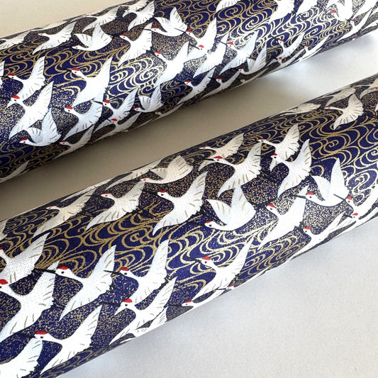 Japanese chiyogami paper with a silkscreened pattern of white cranes in flight on a midnight blue and gold background. Rolled & close-up