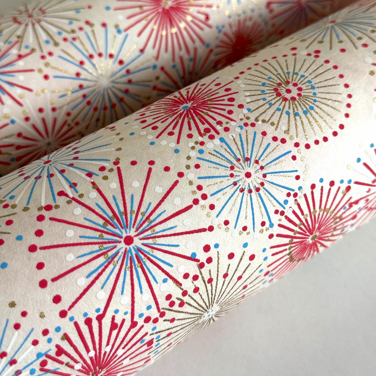 japanese chiyogami silkscreen printed paper with a pattern of fireworks in reds, pinks and lilacs on an ivory background. pictured close-up and rolled