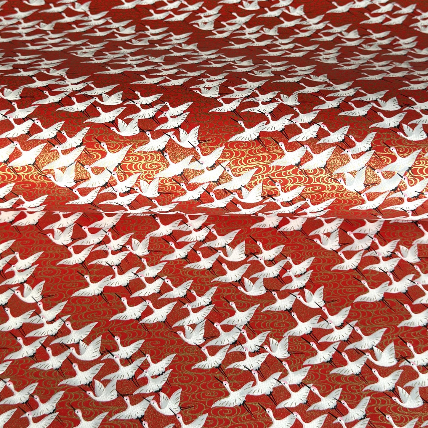A close-up of a red sheet of Japanese chiyogami paper with an all-over repeating pattern of white cranes and small gold wave-like motifs.