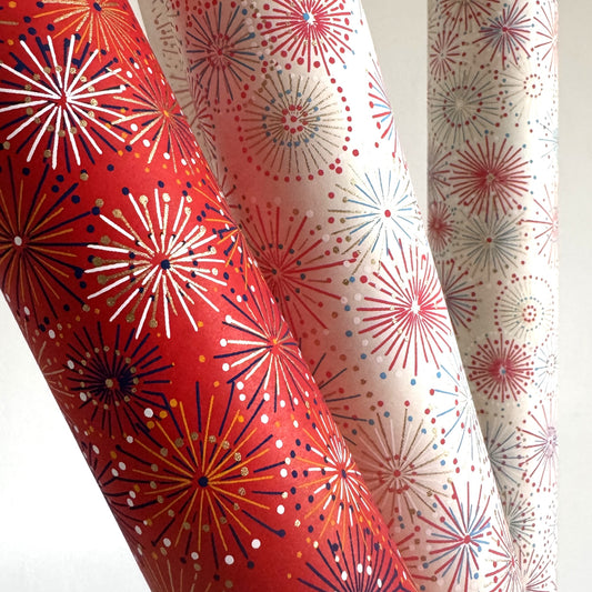 japanese chiyogami silkscreen printed paper with a pattern of fireworks in golds and purples on a red background. Rolled and pictured alongside an ivory version with the same design