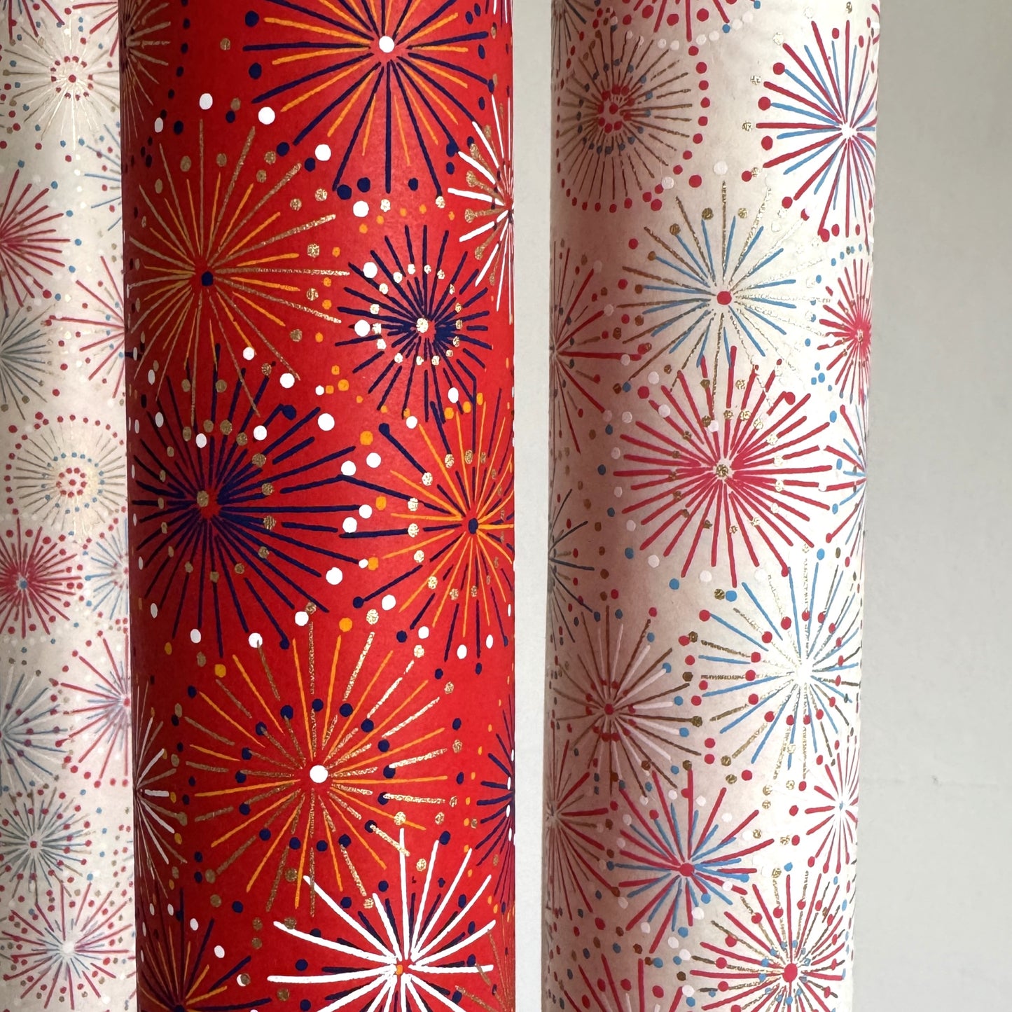 japanese chiyogami silkscreen printed paper with a pattern of fireworks in golds and purples on a red background. Rolled & pictured alongside a version in ivory