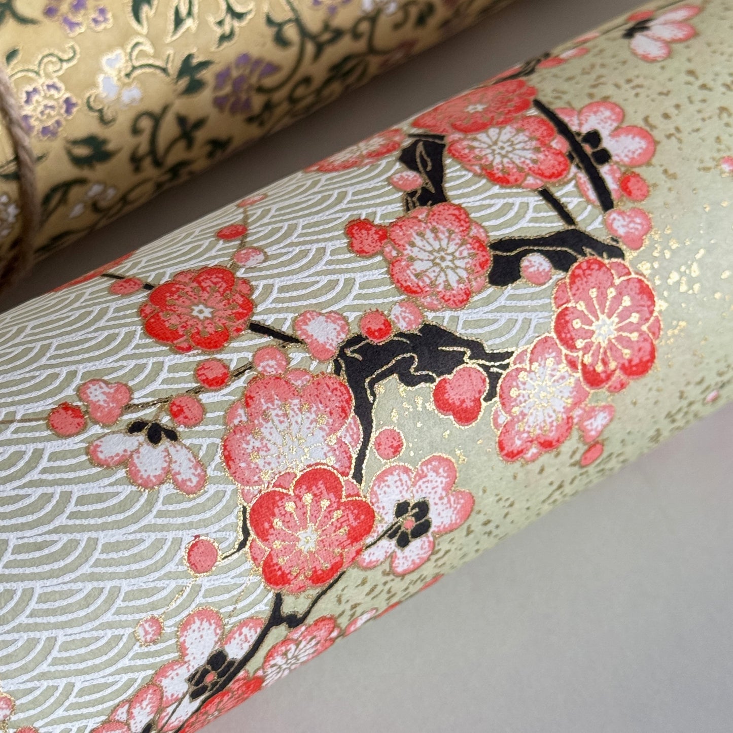 Japanese chiyogami silkscreen paper with diagonal branches of pink plum blossom on a sage green backdrop. Close-up rolled