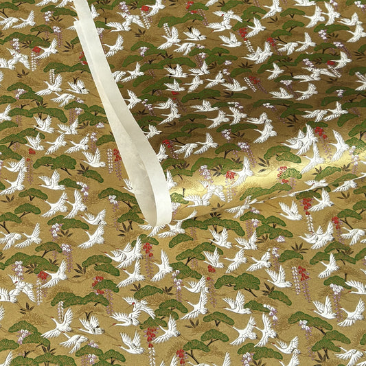 A close-up of a sheet of Japanese chiyogami paper with a repeating Japanese-style pattern featuring white cranes, green pine trees, and small red floral accents on a gold background.