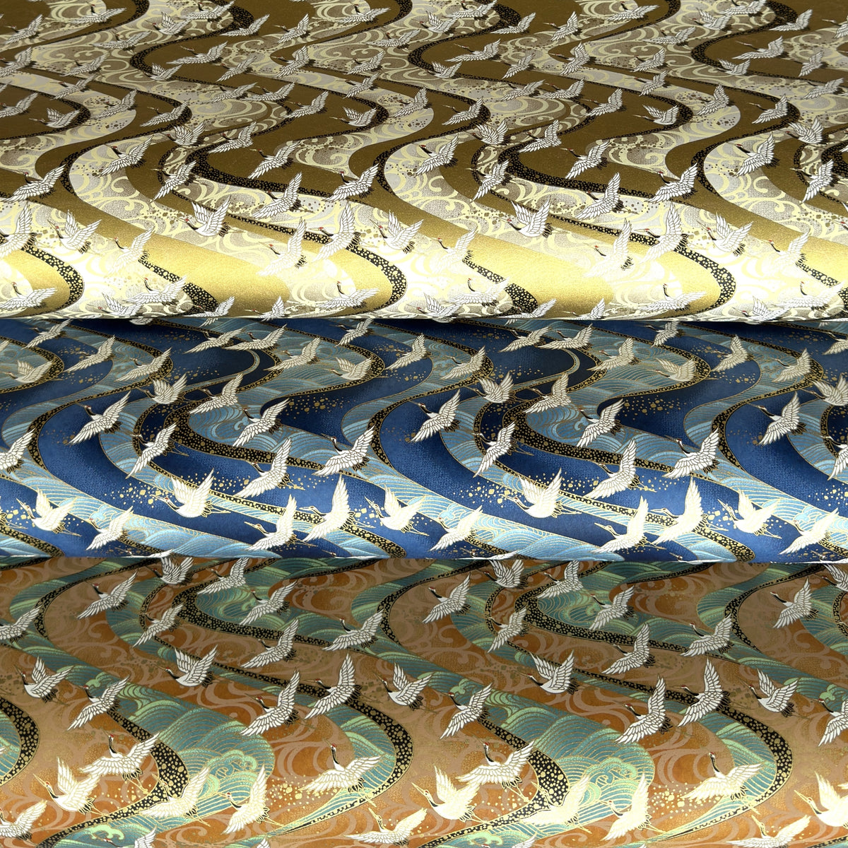three sheets of Japanese chiyogami yuzen paper screen printed with a pattern of white flying cranes on a background of swirling lines. Pictured lightly rolled, top version is gold, middle version is blue and the bottom is tan and aqua