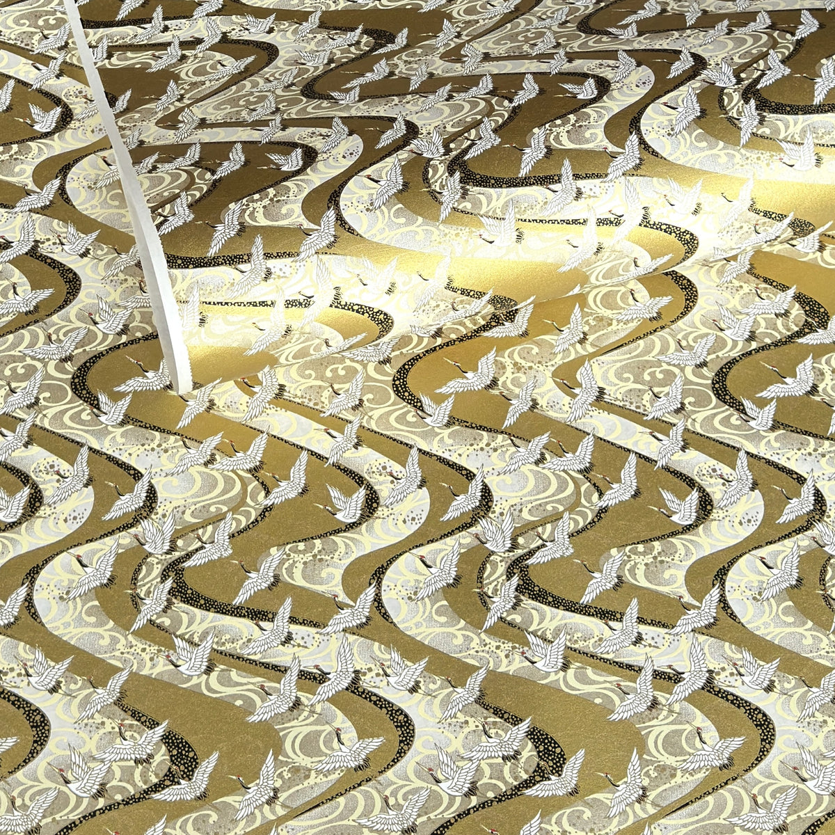 a sheet of Japanese chiyogami paper hand screen printed with a pattern of white cranes in flight. The back drop is silver and lack swirling lines on gold.