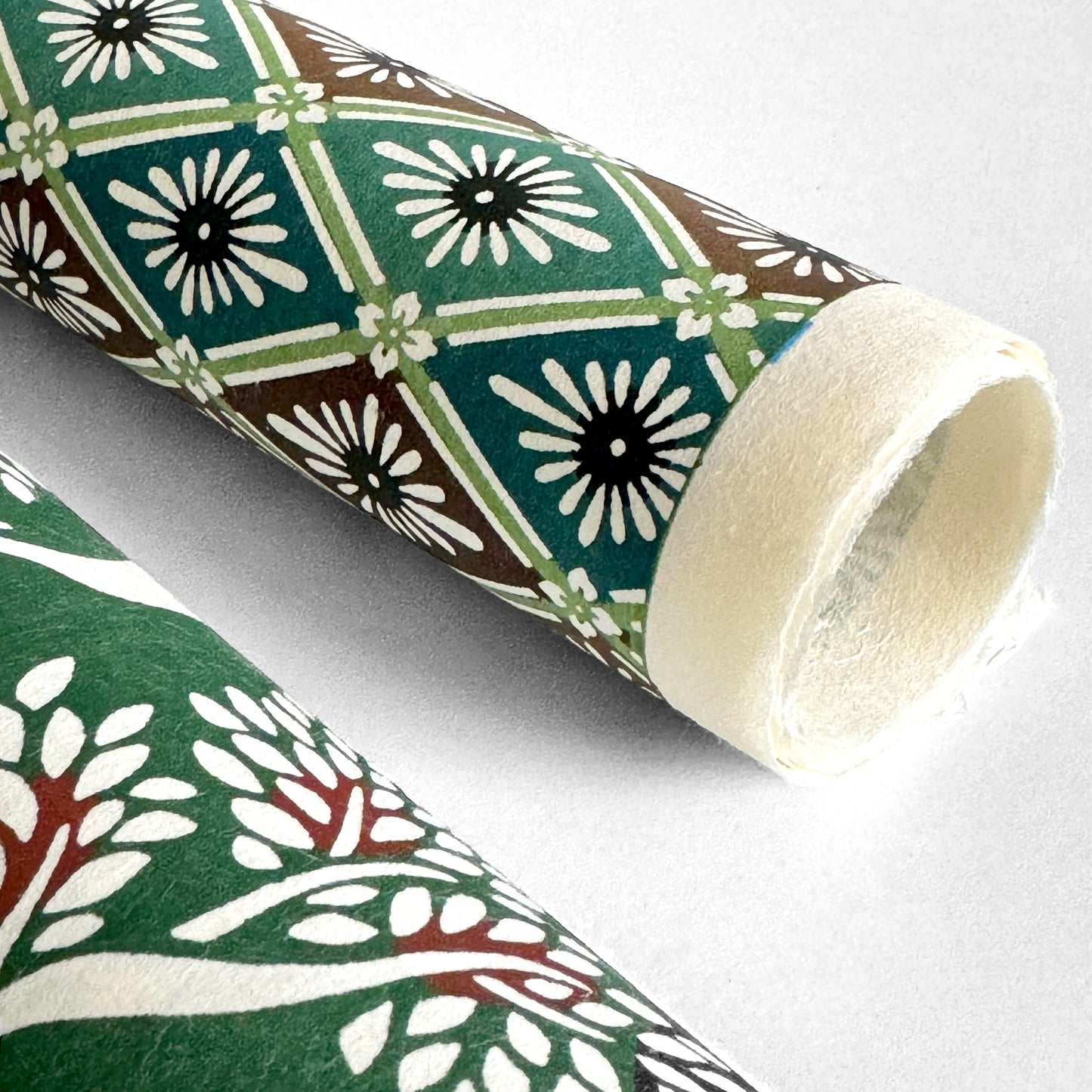 A Japanese stencil-dyed patterned paper with a repeat pattern of floral diamonds in tones of blue, aqua, brown and white. Pictured rolled to show the white deckled edge of the paper