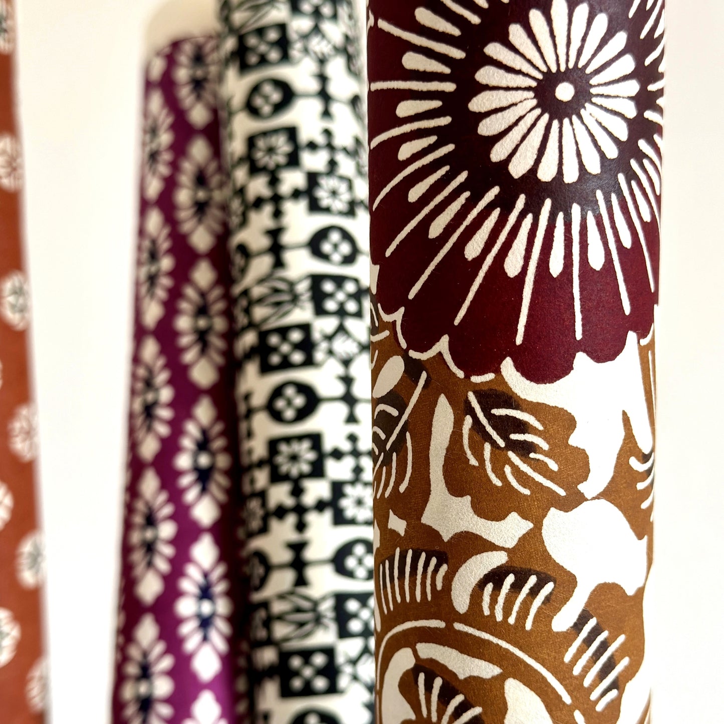 Japanese stencil-dyed paper with a large stylised floral pattern of a chrysanthemum in deep brown and tan.  Close-up, rolled with other papers in the background.