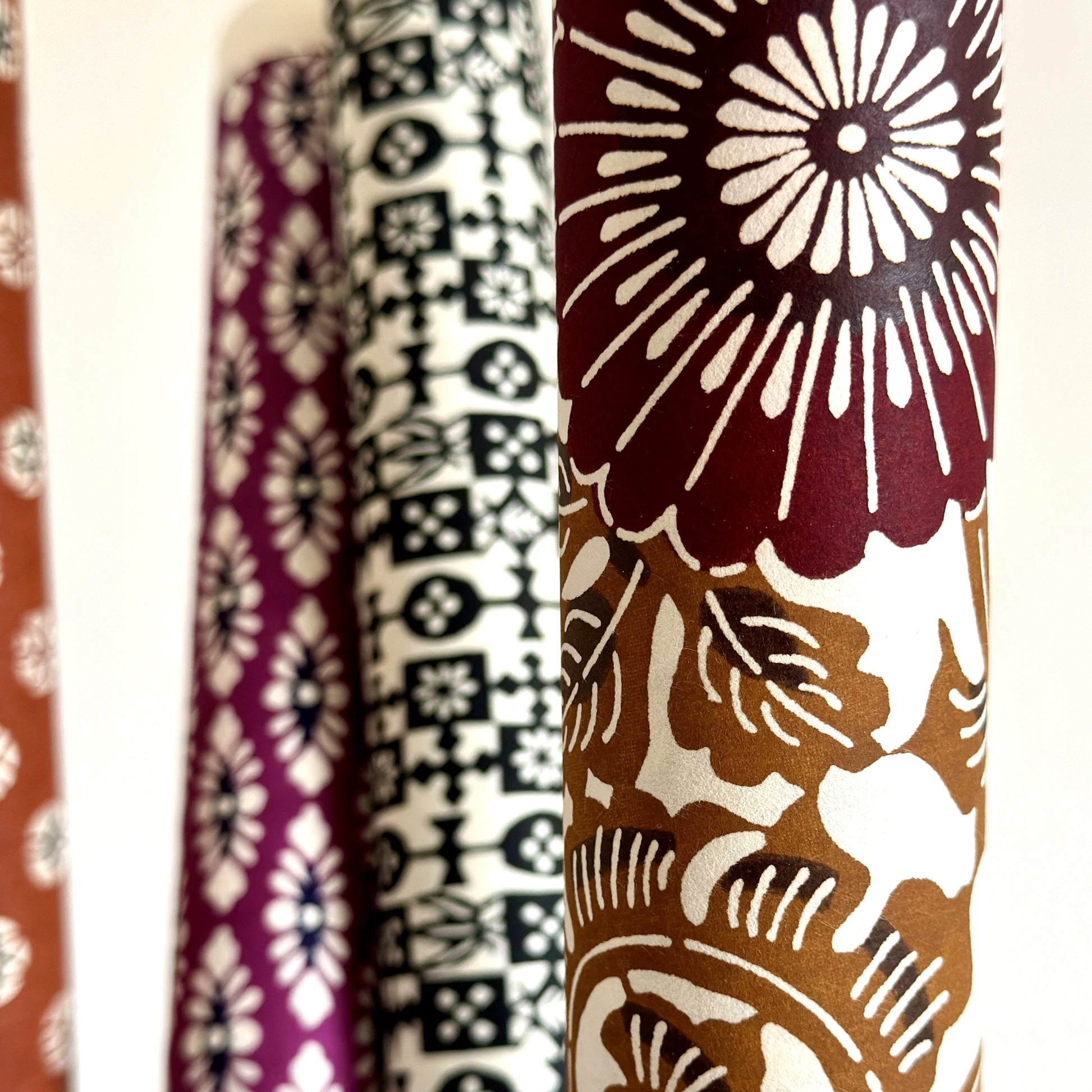 Japanese stencil-dyed paper with a large stylised floral pattern of a chrysanthemum in deep brown and tan.  Close-up, rolled with other papers in the background.