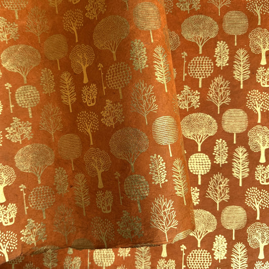 a sheet of screen printed Nepalese lokta paper in terracotta brown with gold trees. The sheet is pictured lightly folded to show the gold of the trees and the deckled edge of the paper.