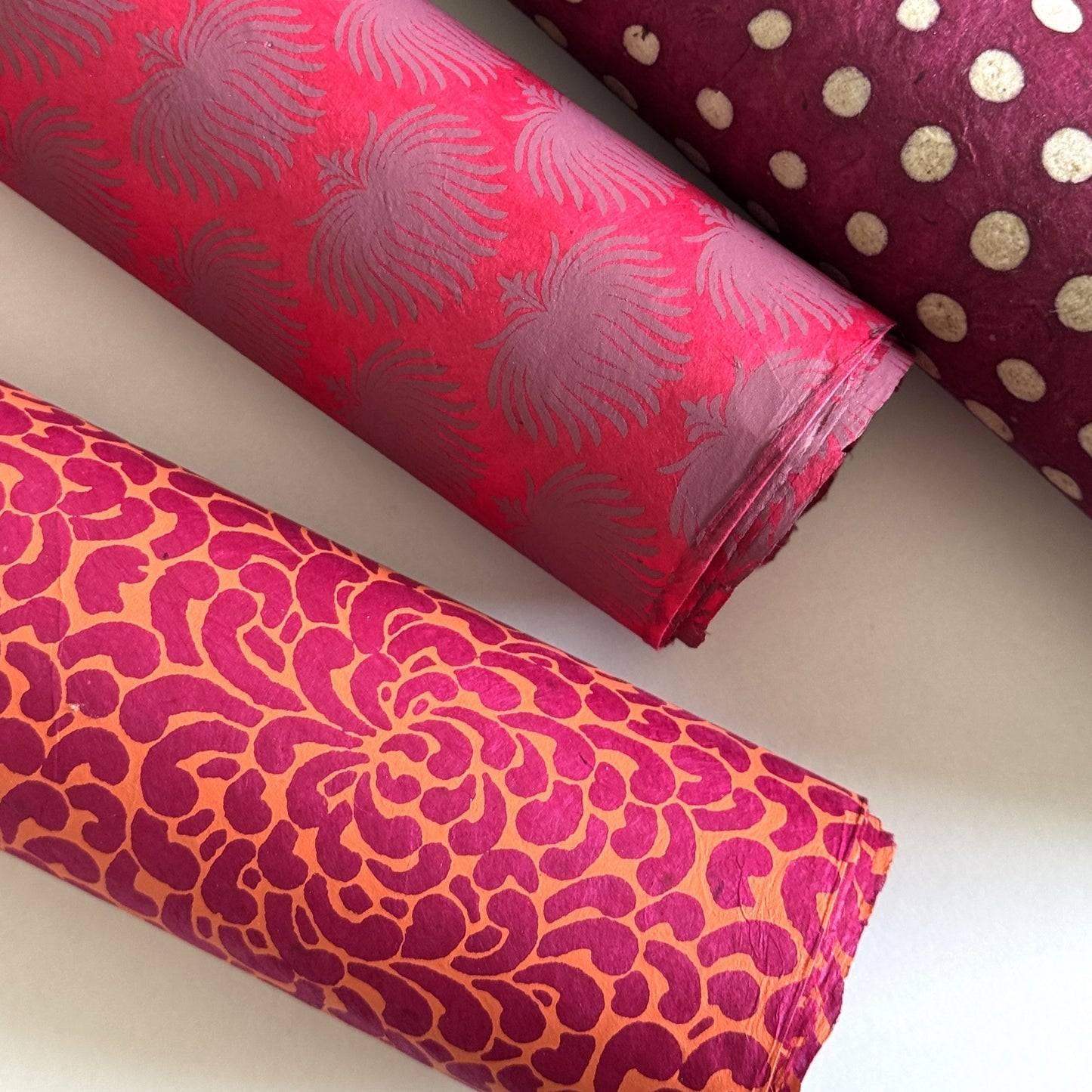 Nepalese handmade lokta paper sheet with a screen printed coral floral design on fuchsia lokta paper. Pictured rolled with other designs