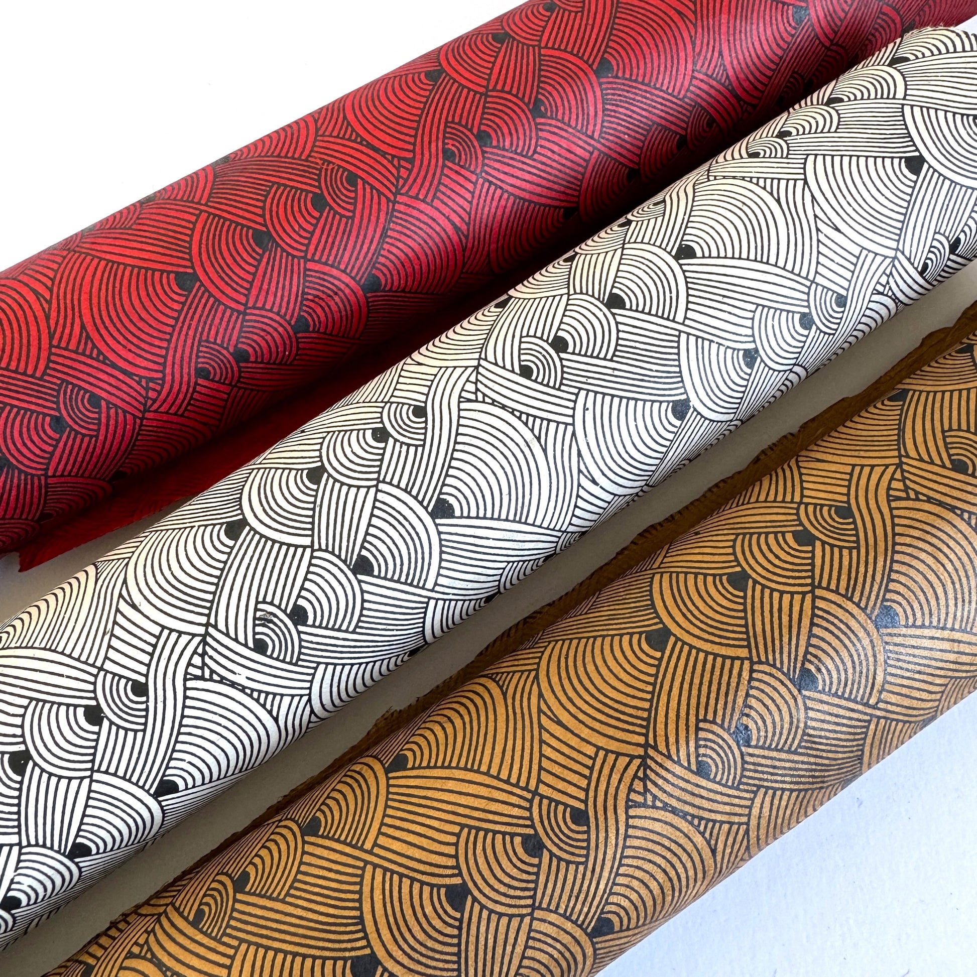 Nepalese handmade decorative lokta paper sheet with a fine lined black knots pattern screen printed on a brown lokta paper. Pictured rolled alongside the same design in different colours