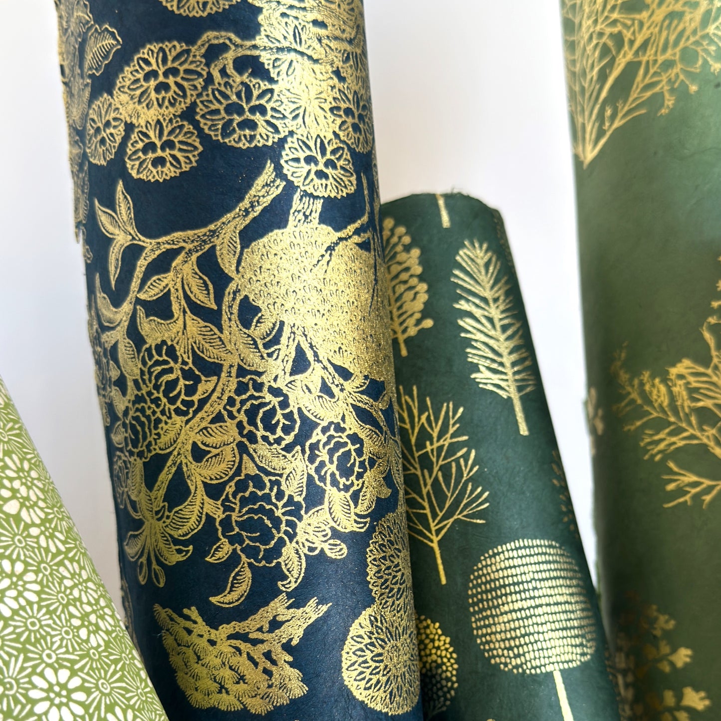 Nepalese handmade decorative lokta paper sheet with a detailed design of trees and flowers in gold metallic screen printed on a midnight blue lokta paper. Rolled alongside other metallic designs
