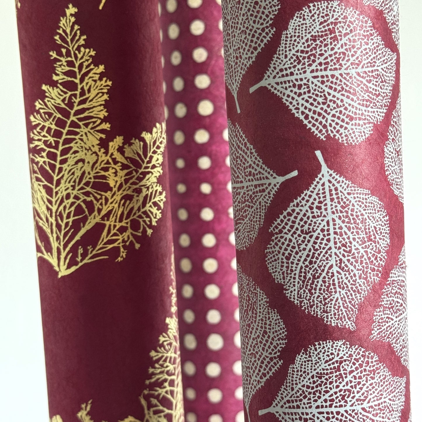 Nepalese handmade decorative lokta paper sheet with a white leaf pattern screen printed on a burgundy lokta paper. Rolled alongside other designs