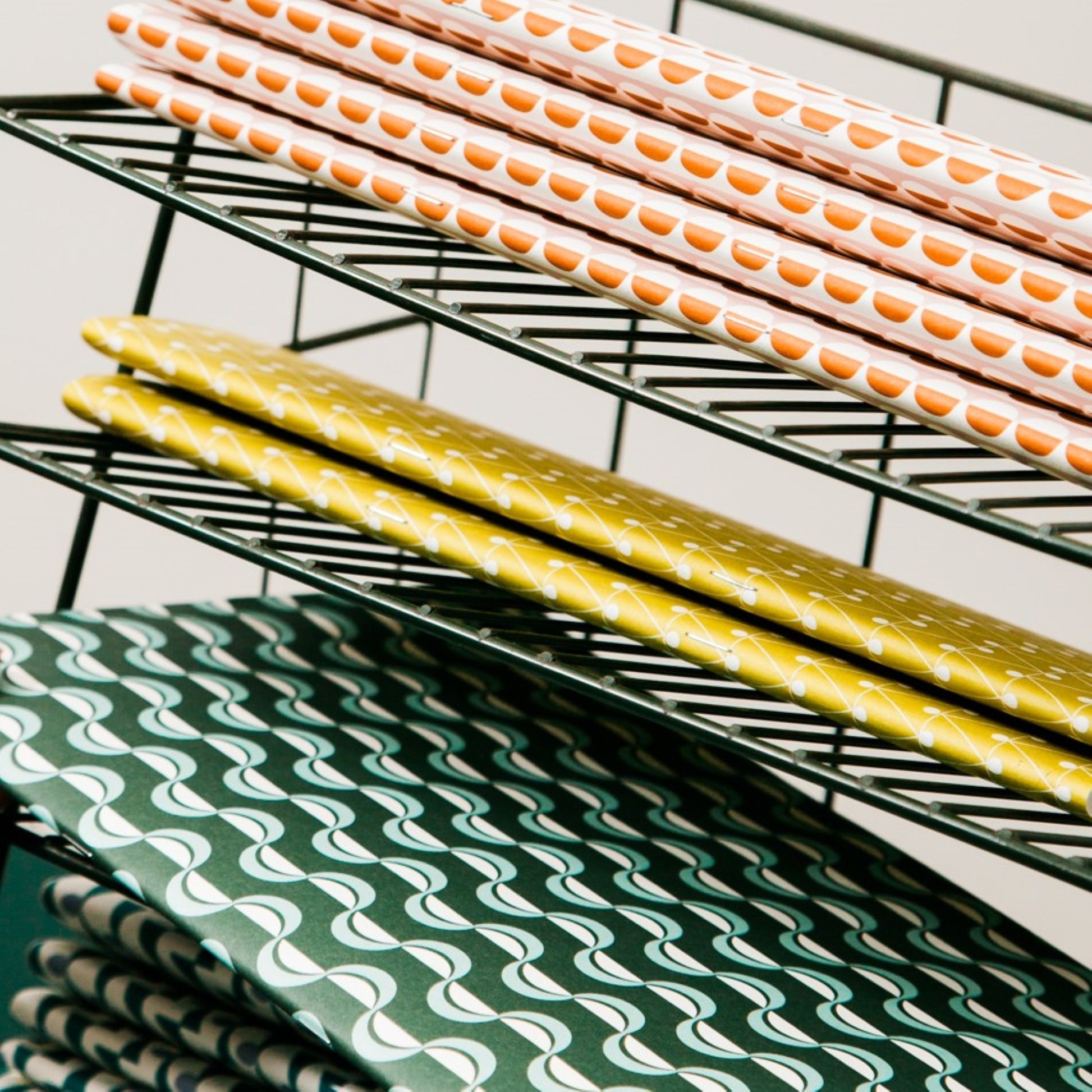 slimline softcover exercise book by Ola Studio, with plain pages. Front cover is a wavy stripe design in dark green and aqua on white. Pictured on a letter rack with other designs from the collection