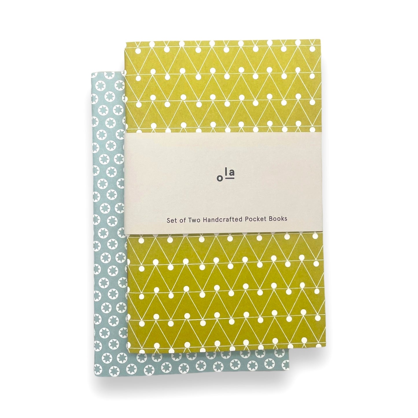 A set of two handcrafted pocket size notebooks by Ola Studio. One cover is a mustard and white triangle geometric design and the other is white stars on an aqua background. Pictured with branded belly band