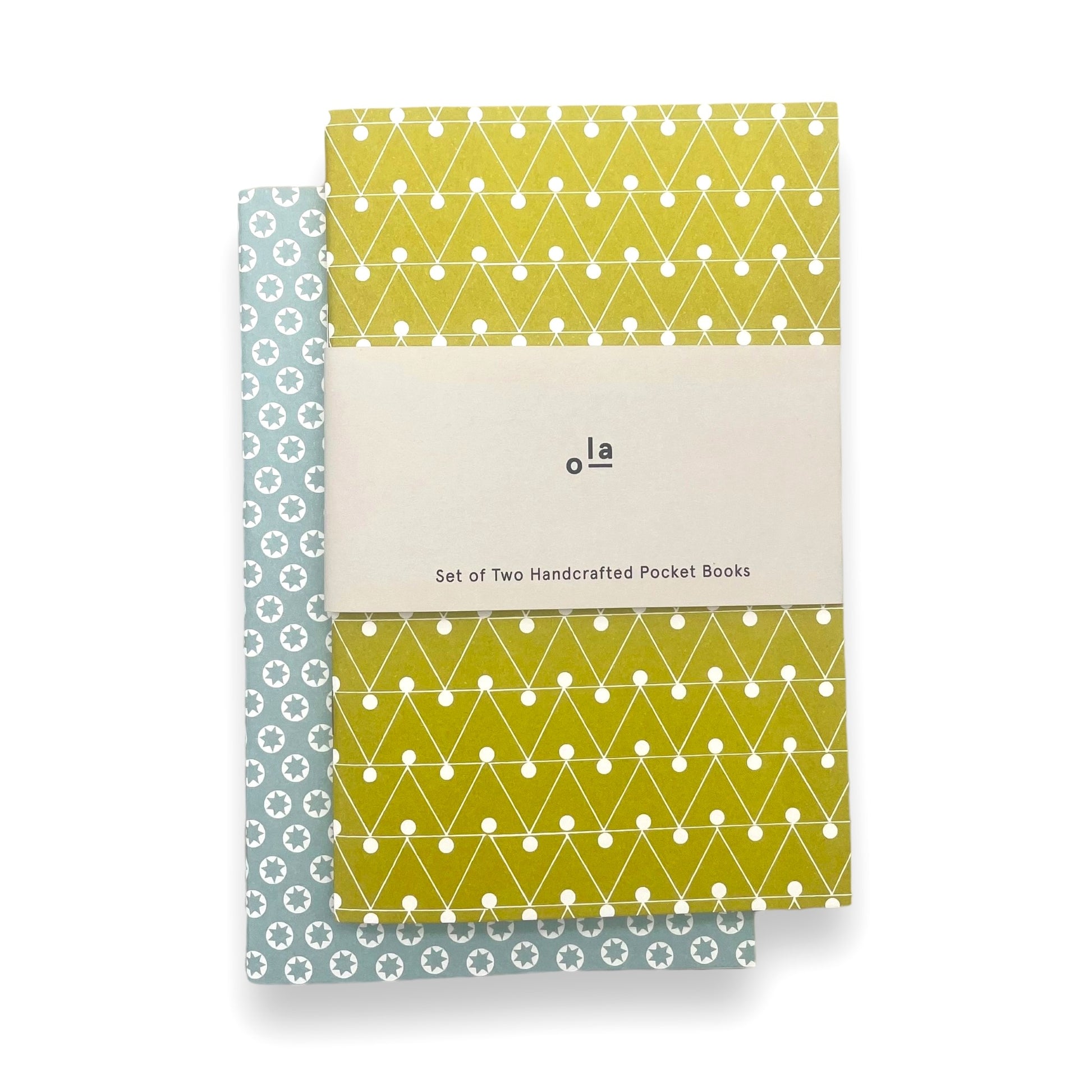A set of two handcrafted pocket size notebooks by Ola Studio. One cover is a mustard and white triangle geometric design and the other is white stars on an aqua background. Pictured with branded belly band