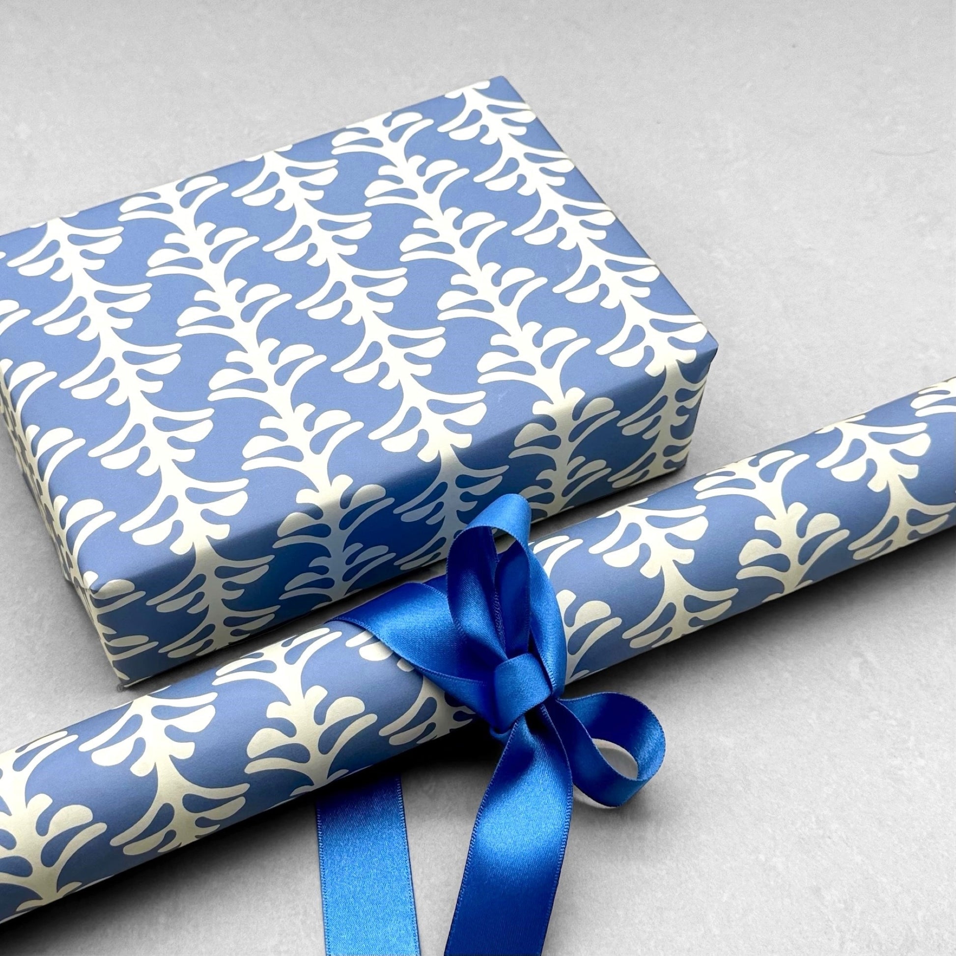 wrapping paper by Otto Editions with a cut-out palm design in white on a sky blue background. Pictured wrapped as a present with a roll of the paper