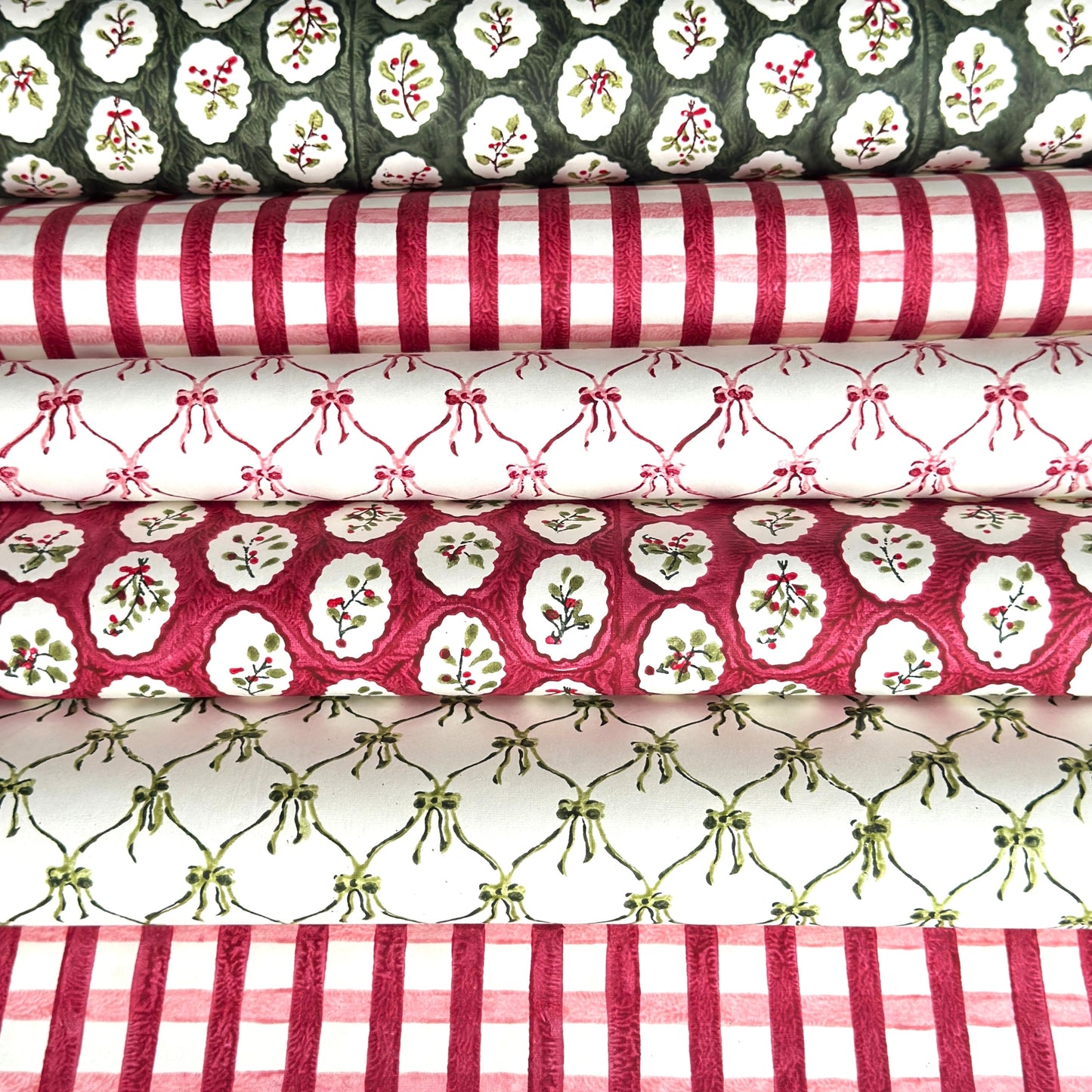 sheets of hand block printed paper in various patterns in red green and wite. there are gingham, small bows and winter foliage patterns