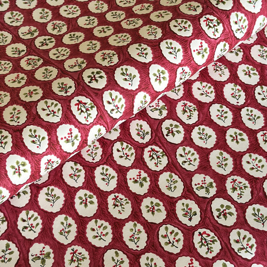 A close-up of a hand block printed paper with a repeating pattern of cream medallions featuring green leaves and red berries on a deep red background.