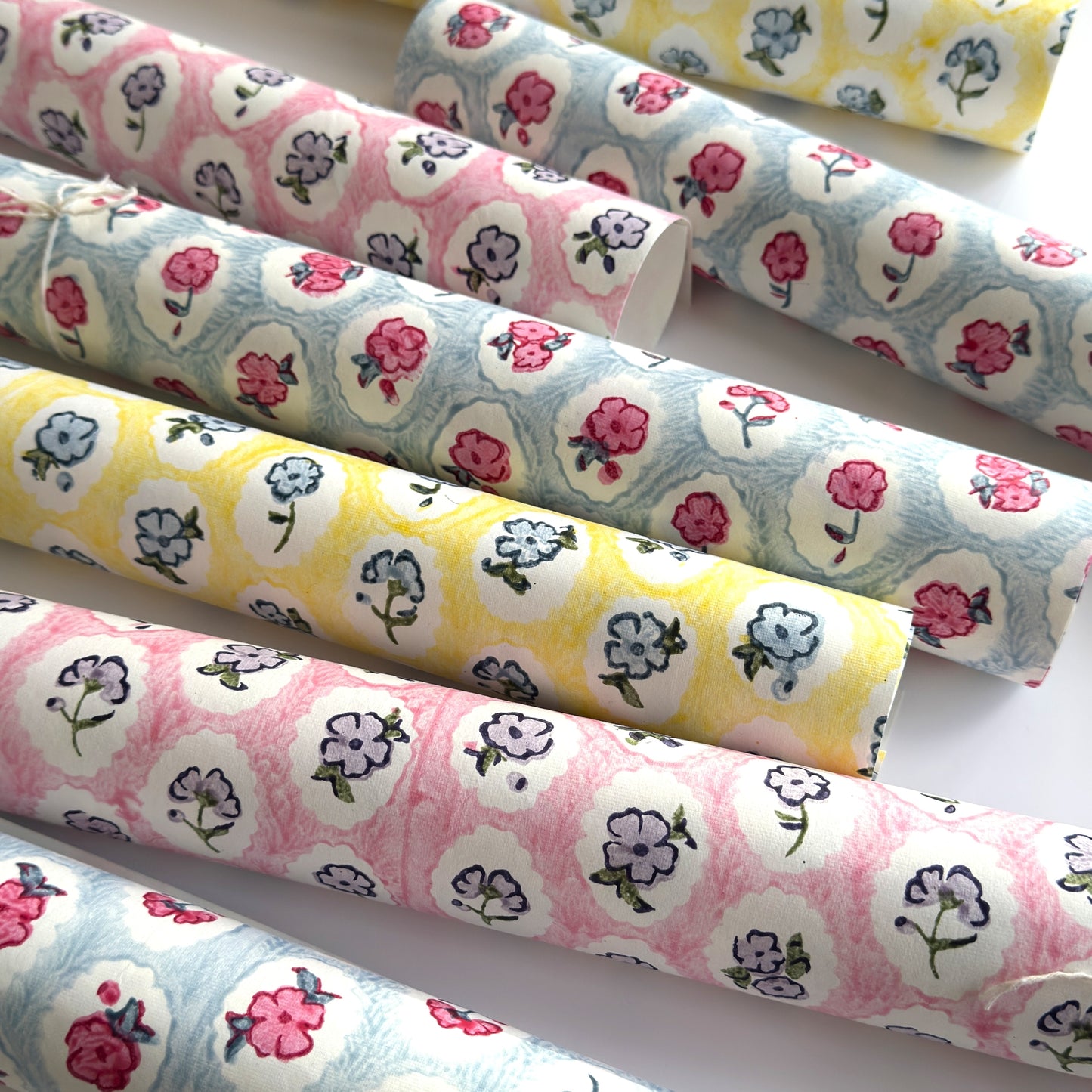 a sheet of hand block printed wrapping paper with a ditsy floral pattern of pink flowers in white ovals with a soft blue backdrop. Pictured rolled with other colour options of the same design