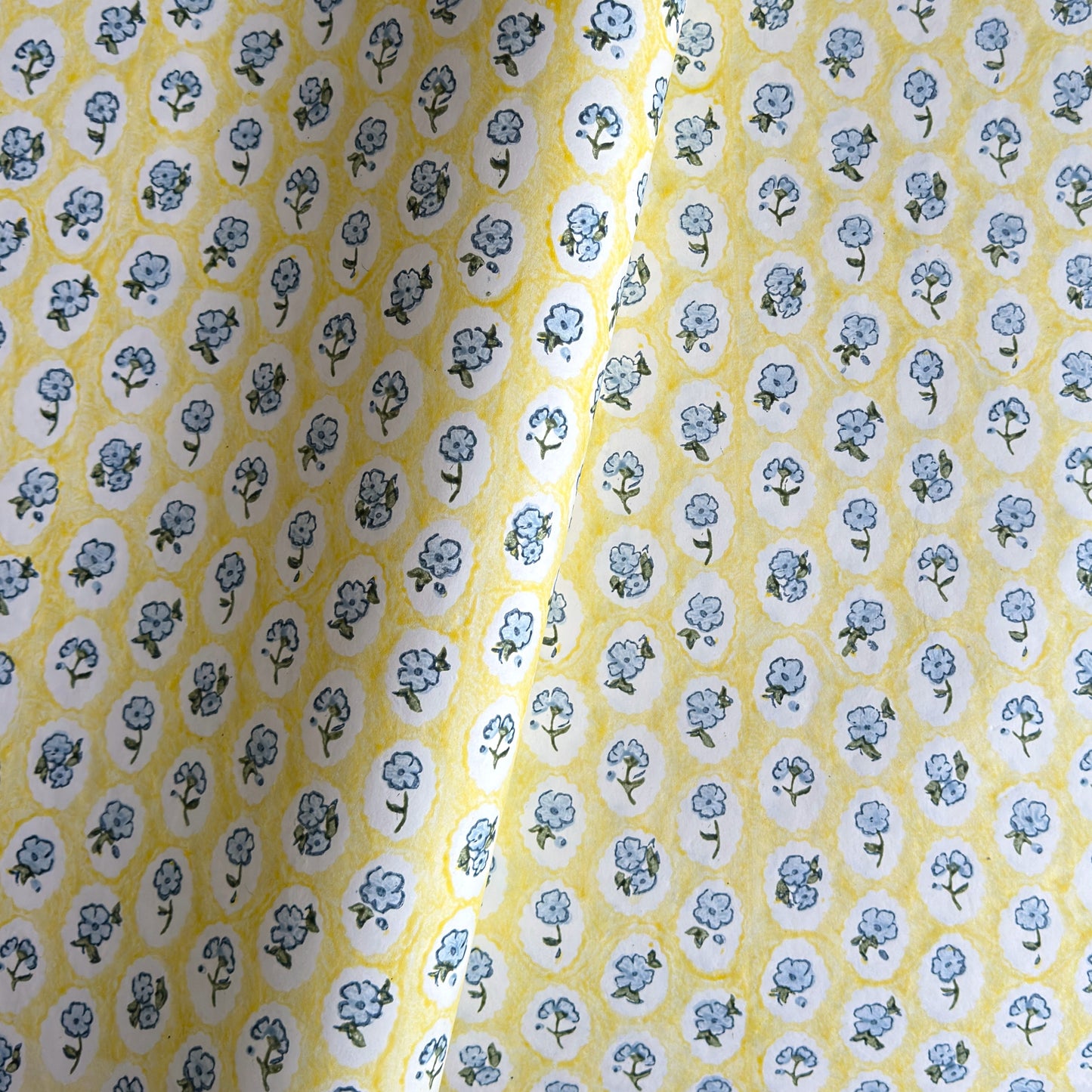 a sheet of hand block printed wrapping paper with a ditsy floral pattern of blue flowers in white ovals with a soft yellow backdrop