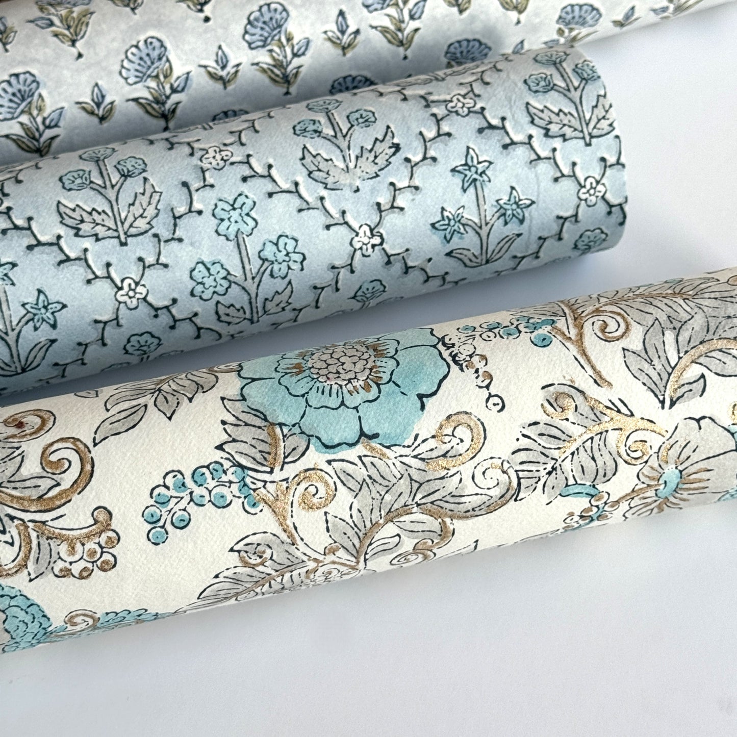 a sheet of hand block printed wrapping paper with a floral wild rose design in opal blue and gold. Pictured rolled with other designs