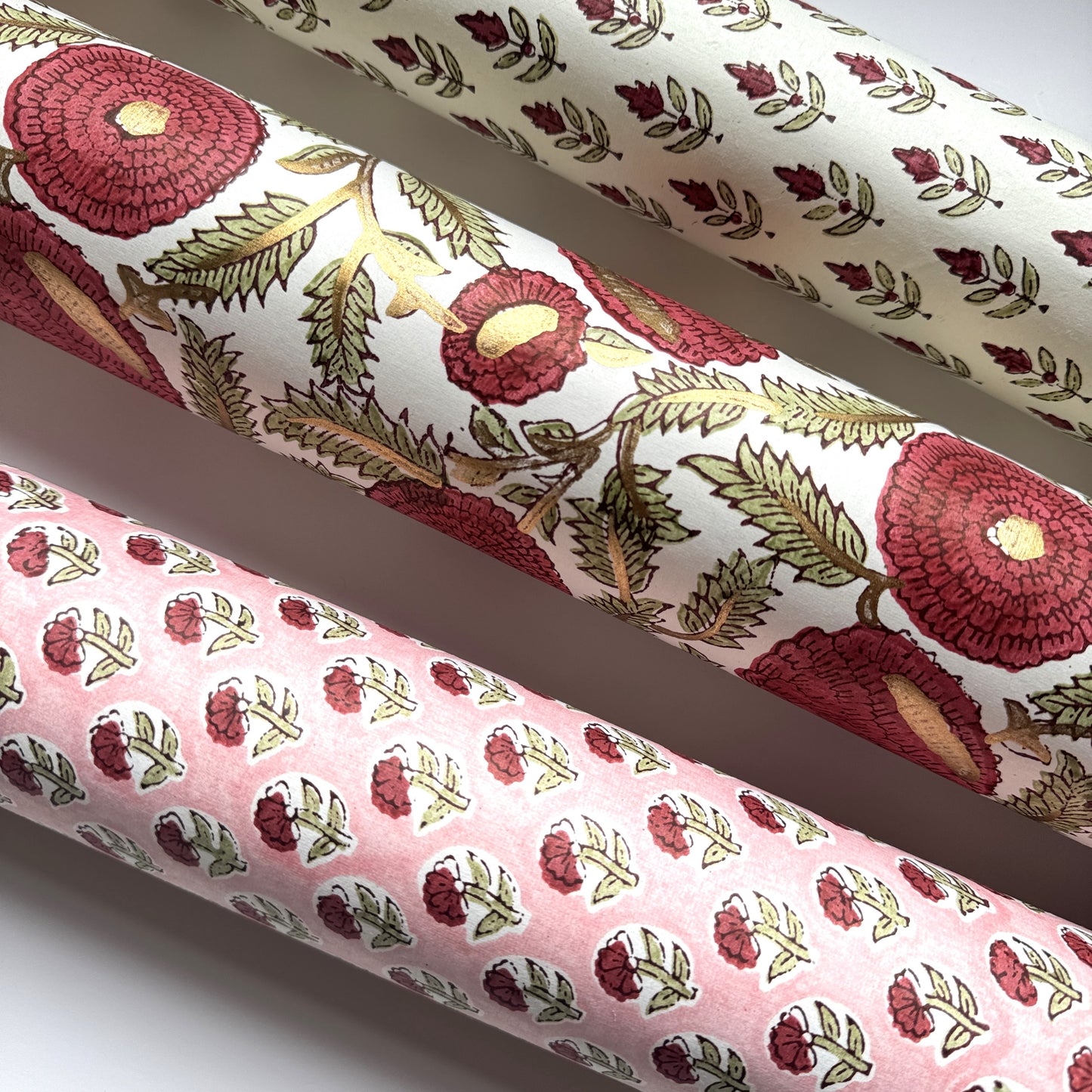block printed paper by Paper Mirchi with a repeat block print pattern of a little stylised flower in dark burgundy and soft green on white backdrop. Pictured rolled with two other coordinating patterns