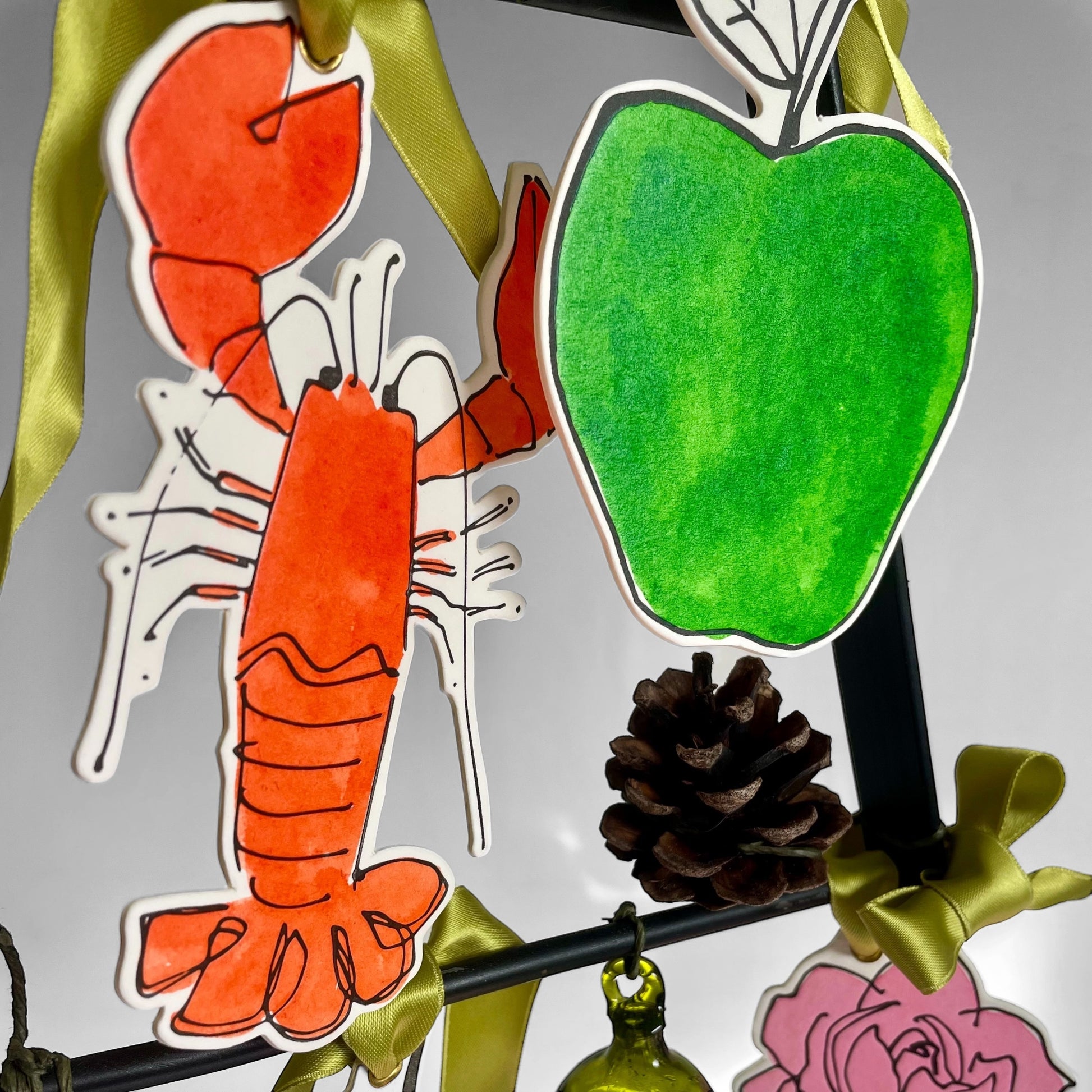 an apple shaped hanging ornament made of thick 1050gsm off-white card, letterpress printed in black and then hand painted in bright green ink. It has a chartreuse green satin ribbon to hang the ornament with. By Scribble & Daub. Hanging on a tree