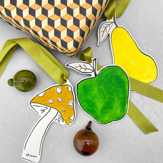 an apple shaped hanging ornament made of thick 1050gsm off-white card, letterpress printed in black and then hand painted in bright green ink. It has a chartreuse green satin ribbon to hang the ornament with. By Scribble & Daub. Pictured being used a a gift tag