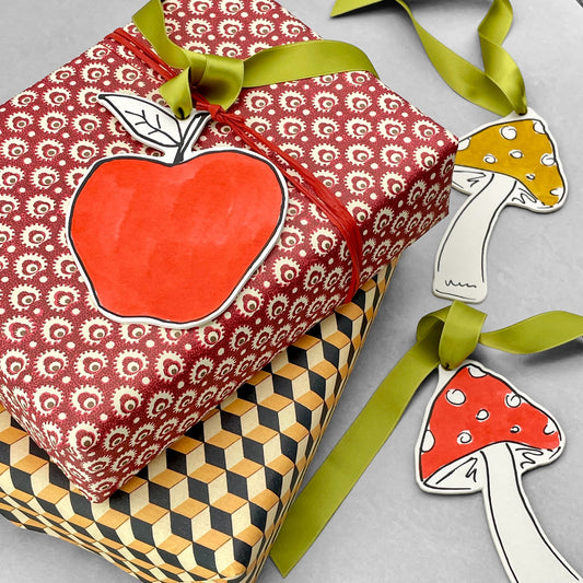 an apple shaped hanging ornament made of thick 1050gsm off-white card, letterpress printed in black and then hand painted in bright red ink. It has a chartreuse green satin ribbon to hang the ornament with. By Scribble & Daub.. Pictured used as a gift tag