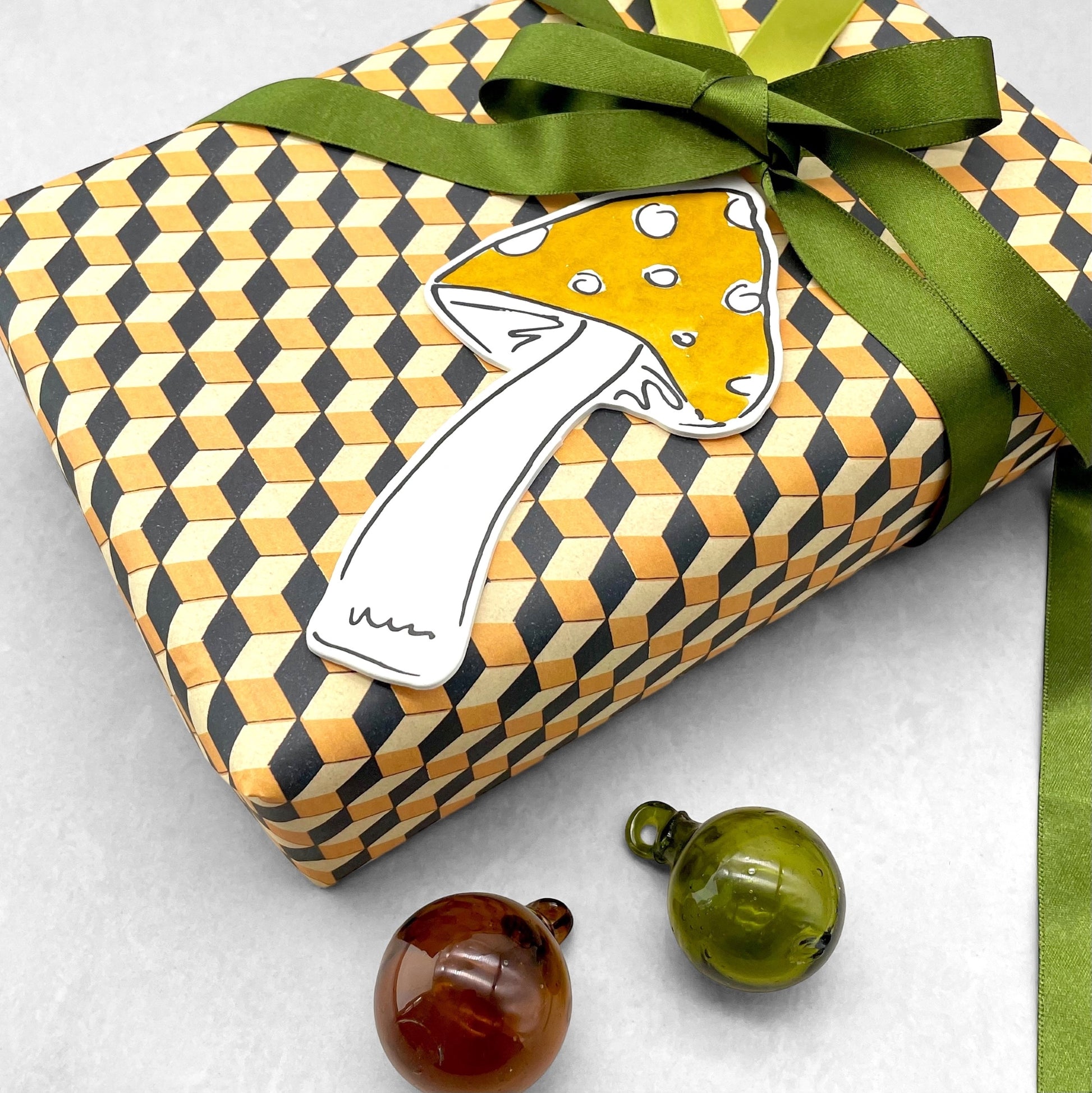 a mushroom shaped hanging ornament made of thick 1050gsm off-white card, letterpress printed in black and then hand painted in ochre ink. It has a chartreuse green satin ribbon to hang the ornament with. By Scribble & Daub. Pictured as a gift tag