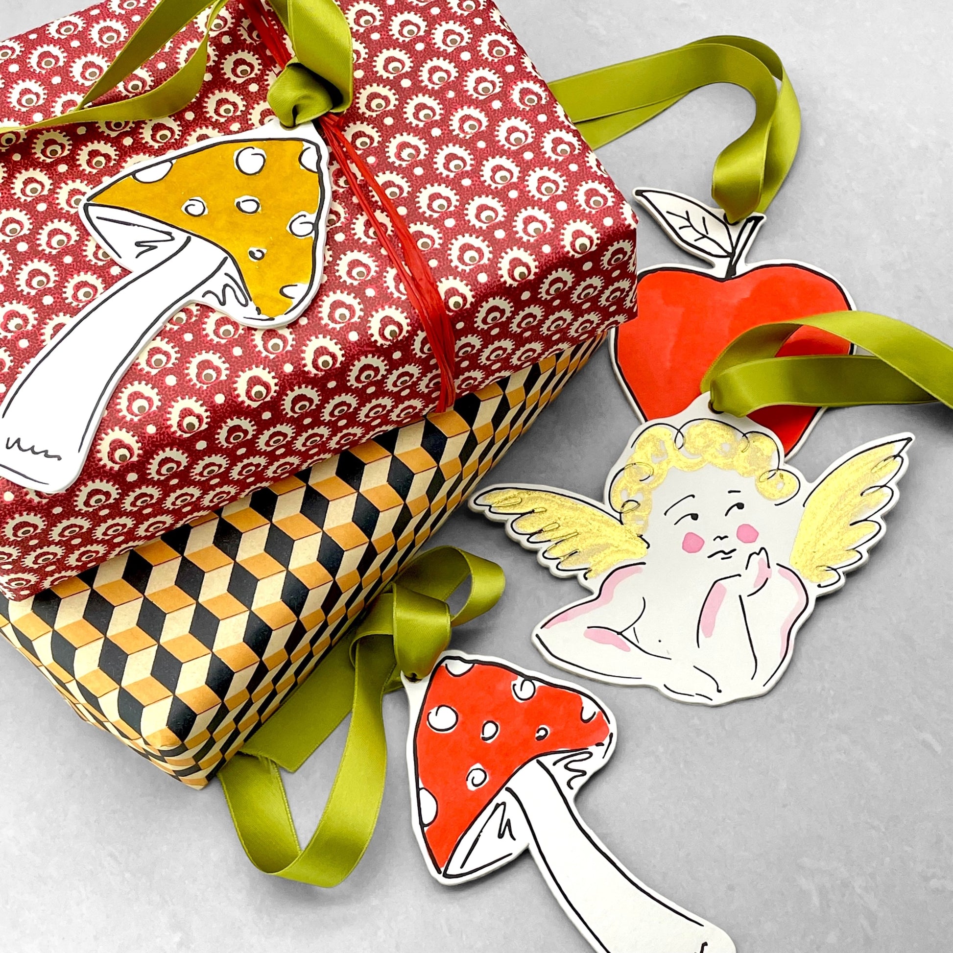 a mushroom shaped hanging ornament made of thick 1050gsm off-white card, letterpress printed in black and then hand painted in red ink. It has a chartreuse green satin ribbon to hang the ornament with. By Scribble & Daub. Pictured as a gift tag