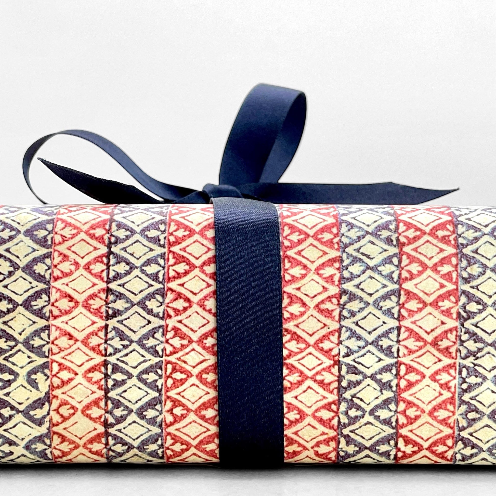 italian Remondini patterned wrapping paper by Tassotti. Striped design with bands red and blue with a cut out diamond effect in cream. Close up with navy ribbon bow